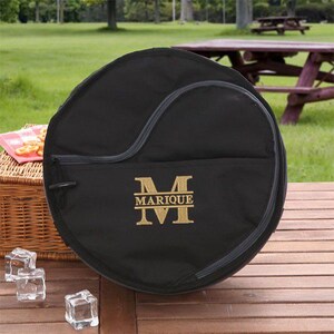 Puede incluir: Bolsa redonda de lona negra con cierre de cremallera y un monograma bordado en oro que dice "MARIQUE" con una "M" grande.