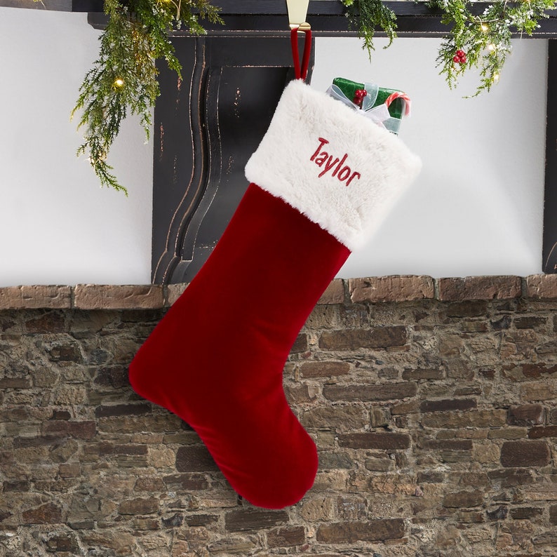 May include: A deep red velvet Christmas stocking featuring a plush white faux fur cuff. The name Taylor is embroidered in red script on the white cuff. A small gift wrapped in green paper with a white ribbon is tucked inside the stocking.