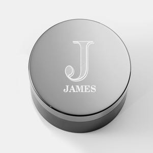 May include: A round, silver-colored container with a black base. The lid is engraved with the letter "J" and the name "JAMES" in a white, elegant font. The container has a reflective, metallic finish.