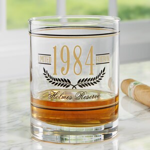 May include: A clear glass tumbler with gold accents and the text "1984 Limited Edition" and "Holmes Reserve". The glass is partially filled with amber liquid. A cigar rests on the side.