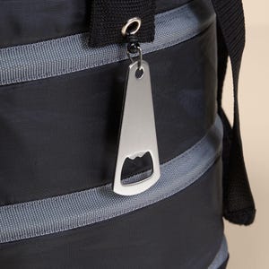 Puede incluir: Una bolsa nevera negra y gris con un abridor de botellas plateado sujeto al tirador de la cremallera. La bolsa tiene una cremallera y una correa para el hombro.