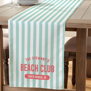 May include: A striped table runner with alternating white and light blue vertical stripes. The text "THE STEWART'S BEACH CLUB SANTA MONICA, CA" is printed in red and white on the lower portion of the runner. The runner is on a wooden table.