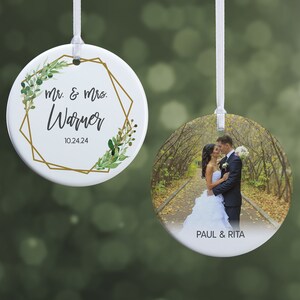 May include: Two white ceramic ornaments with white ribbons. One features a photo of a couple kissing, with the text "PAUL & RITA". The other has the text "Mr. & Mrs. Warner" and the date "10.24.24" inside a gold geometric frame with green leaves.