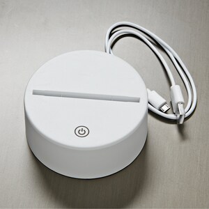 May include: A white, circular LED light base with a slot for an acrylic insert. It features a power button icon and includes a white USB cable for power. The base is designed to illuminate decorative acrylic pieces.