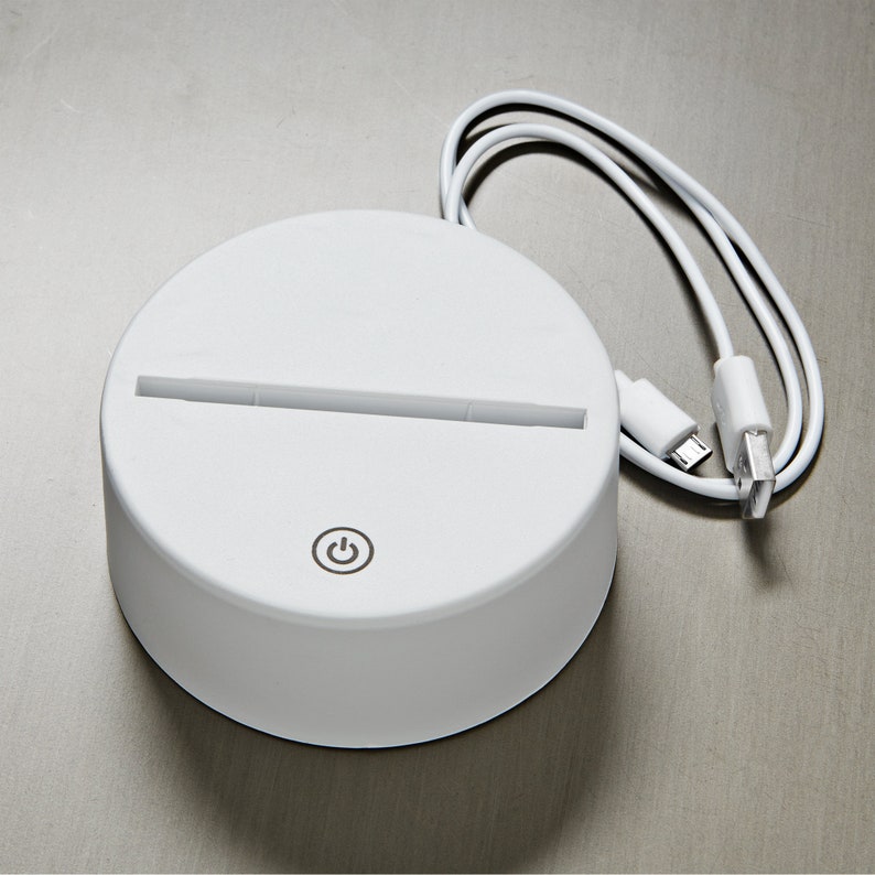 May include: A white, circular LED light base with a slot for an acrylic insert. It features a power button icon and includes a white USB cable for power. The base is designed to illuminate decorative acrylic pieces.
