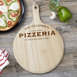 May include: A round wooden pizza serving board with a handle and a leather hanging loop. The board is engraved with the text THE CALLAHAN'S oven-baked PIZZERIA the best frozen pizza in town! The wood has a light natural grain and smooth finish.