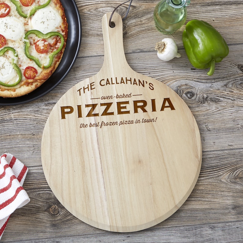 May include: A round wooden pizza serving board with a handle and a leather hanging loop. The board is engraved with the text THE CALLAHAN'S oven-baked PIZZERIA the best frozen pizza in town! The wood has a light natural grain and smooth finish.