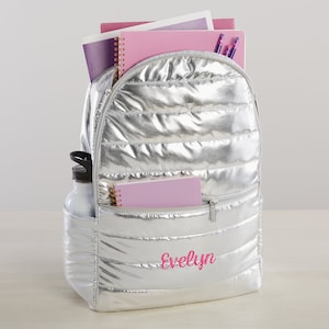 May include: Silver quilted backpack with a pink zipper and the name "Evelyn" embroidered on the front. The backpack is filled with school supplies, including notebooks and pens.