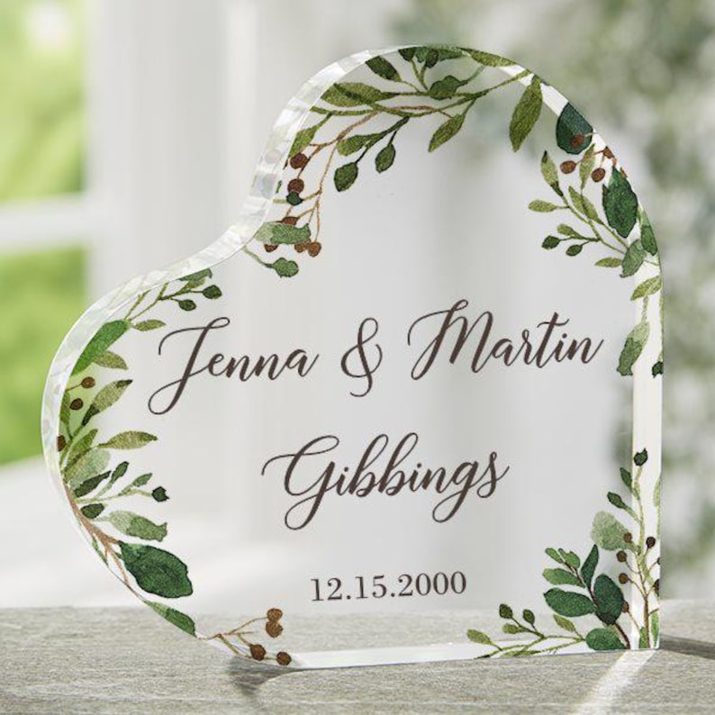 May include: A clear heart-shaped glass keepsake with a leafy green and brown floral design. The text "Jenna & Martin Gibbing 12.15.2000" is printed on the glass.