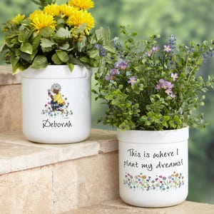May include: Two white ceramic flower pots with colorful floral designs. One pot features a woman gardening with the name "Deborah" written below. The other pot has the text "This is where I plant my dreams!"