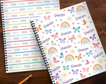 May include: Two spiral bound notebooks with different designs. The first notebook has a white background with the name Amelia repeated in a colorful pattern. The second notebook has a white background with a colorful rainbow, butterflies, and the name Vanessa repeated in a colorful pattern.