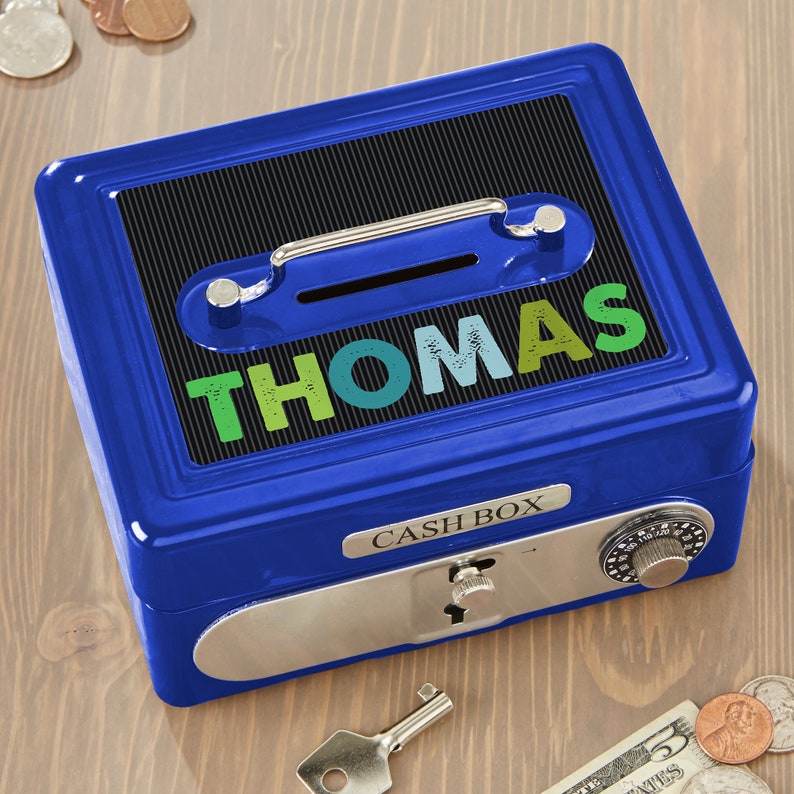 Puede incluir: Una caja de efectivo met&aacute;lica azul con el nombre "THOMAS" en letras coloridas. La caja tiene una cerradura de combinaci&oacute;n, una ranura para monedas y un asa. Se ve una llave y algunas monedas.