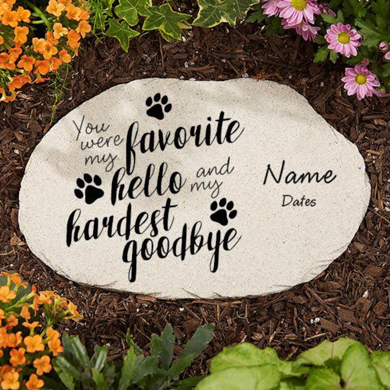May include: A light-colored stone garden marker with the words "You were my favorite hello and my hardest goodbye." Paw prints and space for a name and dates are also included. The stone is surrounded by flowers and mulch.