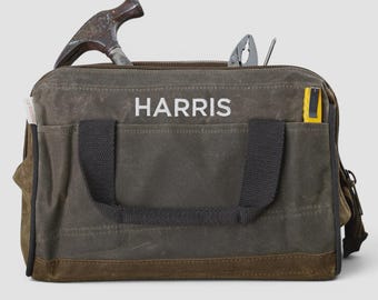 May include: A dark olive green canvas tool bag with black fabric handles and a brown leather base. The word HARRIS is printed in white on the front. A metal hammer, pliers, and a yellow utility knife are tucked inside the open top compartment.
