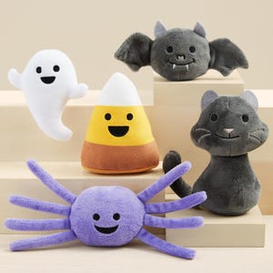 K&ouml;nnte beinhalten: Eine Sammlung von Pl&uuml;schspielzeugen zum Thema Halloween, darunter ein wei&szlig;es Gespenst, ein Maisbonbon, eine graue Fledermaus, eine graue Katze und eine lila Spinne. Jedes Spielzeug hat ein l&auml;chelndes Gesicht und ist aus weichem Stoff.