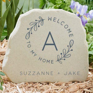 May include: A beige stone garden plaque with the text "WELCOME TO MY HOME" in a circular design around the letter "A". The names "SUZZANNE + JAKE" are printed at the bottom. The plaque is surrounded by wood chips and greenery.