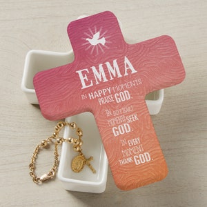 May include: A white ceramic cross-shaped box with a pink and orange ombre cross lid. The lid has the name "EMMA" and the text "IN HAPPY MOMENTS PRAISE GOD. IN DIFFICULT MOMENTS SEEK GOD. IN EVERY MOMENT THANK GOD."