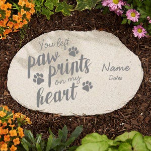 May include: A light-colored stone garden marker with the text "You left paw prints on my heart" and paw print graphics. The marker also includes spaces for a name and dates. The stone is surrounded by mulch and flowers.