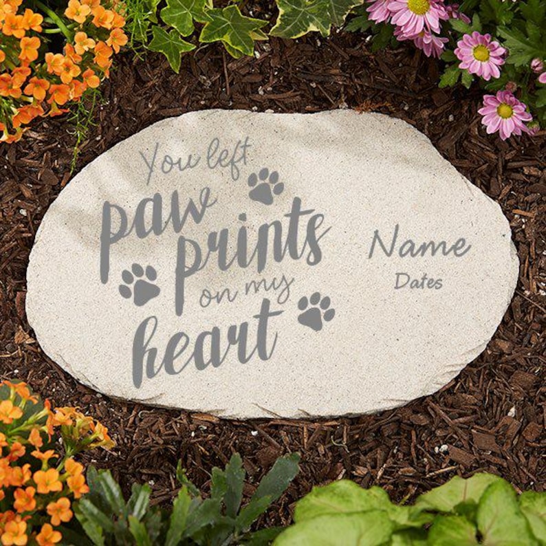 May include: A light-colored stone garden marker with the text "You left paw prints on my heart" and paw print graphics. The marker also includes spaces for a name and dates. The stone is surrounded by mulch and flowers.