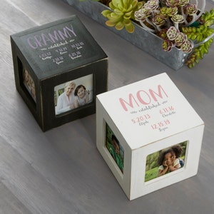 May include: Two wooden photo cube boxes with a white and black finish. The boxes have a space for four photos and are personalized with the words "GRAMMY" and "MOM" along with dates and names.