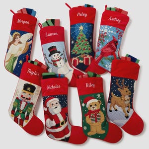 May include: Eight red Christmas stockings featuring embroidered names and festive holiday imagery including an angel, snowman, Christmas tree, cardinal, nutcracker, Santa Claus, teddy bear, and reindeer, each filled with small rectangular gift boxes.