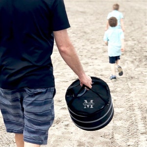 Puede incluir: Una bolsa de enfriador cil&iacute;ndrica negra y gris con cierre de cremallera y asa. La bolsa tiene una franja blanca alrededor del medio y las iniciales "M.M." bordadas en la parte delantera. La bolsa la lleva una persona que lleva una camisa negra y pantalones cortos grises.