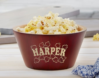 May include: A wooden popcorn bowl filled with popcorn. The bowl is a deep red color with a lighter wood rim. The name "HARPER" is printed on the front in a cream-colored font, surrounded by popcorn graphics. The bowl is sitting on a white wooden surface.