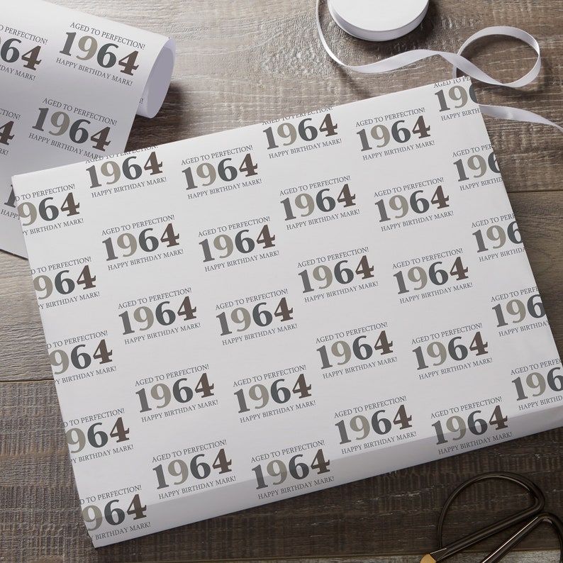 May include: White wrapping paper with a repeating pattern of the year "1964" in gray and brown, with the text "Aged to Perfection! Happy Birthday Mark!" A roll of paper and white ribbon are visible.