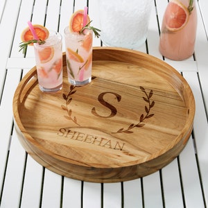 May include: A round wooden serving tray with handles, engraved with the letter "S" inside a laurel wreath and the name "SHEEHAN". Two cocktails with grapefruit slices and rosemary sprigs sit on the tray. A pitcher and a glass of ice are in the background.