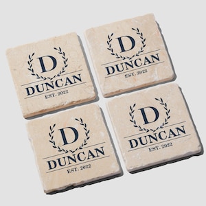 May include: A set of four square, light beige stone coasters featuring a dark blue printed design. Each coaster displays a central letter D enclosed in a laurel wreath, with the text DUNCAN and EST. 2022 printed in a serif font below the wreath.