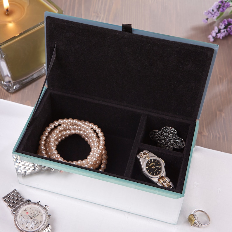 May include: A mirrored jewelry box with a black velvet interior. The box has three compartments and is open to reveal a pearl necklace, a watch, and a decorative pin.