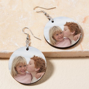 May include: A pair of round wooden earrings with a photo of two children, a girl and a boy, printed on them. The girl is wearing a pink shirt and the boy is wearing a white shirt. The earrings are silver and have a hook closure.