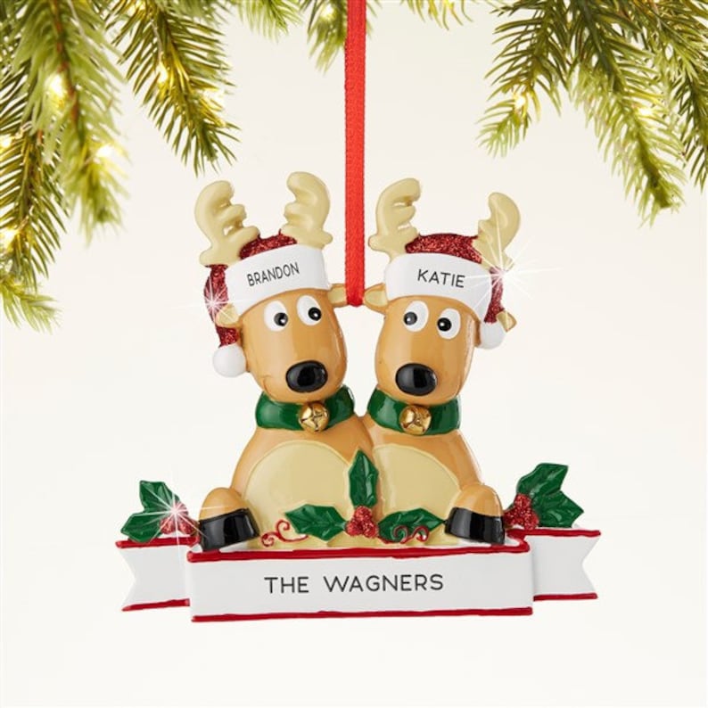 K&ouml;nnte beinhalten: Weihnachtsschmuck mit zwei Rentieren mit Weihnachtsm&uuml;tzen mit den Namen "Brandon" und "Katie". Die Rentiere sind hellbraun mit schwarzen Nasen, gr&uuml;nen Schals und goldenen Glocken. Auf der Basis steht "THE WAGNERS".