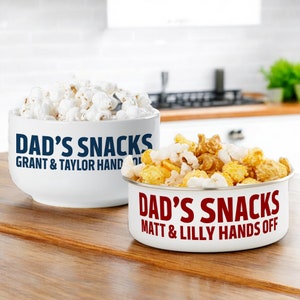May include: Two white ceramic bowls with blue and red text. The bowls are filled with popcorn and a mix of popcorn and caramel corn. The text on the bowls reads "Dad's Snacks" with names underneath. The first bowl says "Grant & Taylor Hands Off" and the second bowl says "Matt & Lilly Hands Off".