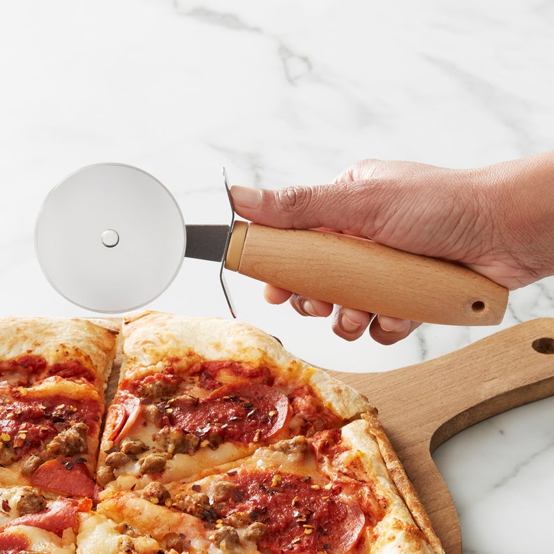 Peut inclure: Un coupe-pizza avec une poign&eacute;e en bois et une lame en acier inoxydable. Le coupe-pizza est utilis&eacute; pour couper une pizza avec du pepperoni, de la saucisse et des flocons de piment rouge.