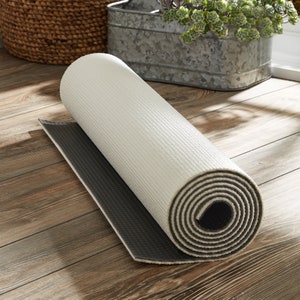 May include: A rolled-up white yoga mat with a black non-slip bottom. The mat is rolled up tightly and is lying on a wooden floor.