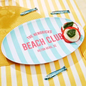 May include: An oval serving platter with blue and white stripes, featuring the text "THE HENDRICKS BEACH CLUB, HILTON HEAD, SC" in pink. The platter holds two tomato and mozzarella appetizers, and is surrounded by decorative fish.