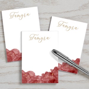 May include: Three white notepads with the name "Tamyra" written in gold script. The notepads have a red and gold watercolor design at the bottom.