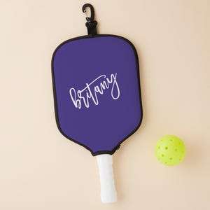 May include: Purple pickleball paddle with a black strap and the name "Brittany" written in white cursive. A yellow pickleball is in the background.