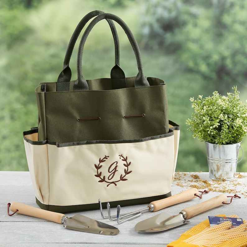 Peut inclure: Un sac de jardinage en toile vert et cr&egrave;me avec une couronne brod&eacute;e marron et la lettre "G" au centre. Le sac est rempli d'outils de jardinage, dont une truelle, un r&acirc;teau &agrave; main et un cultivateur.