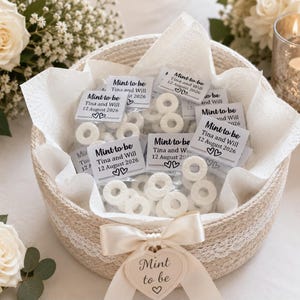 May include: A round woven basket with a white lace trim and a cream ribbon tied in a bow, featuring a heart-shaped tag labelled Mint to be. Inside are clear plastic bags of white ring-shaped mints with labels reading Mint to be, Tina and Will, 12 August 2026.