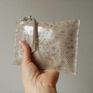 May include: A rectangular clutch with a snakeskin pattern in shades of beige and brown. The clutch has a small loop and a matching strap. The bag is held in a hand.