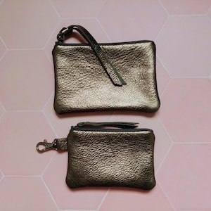 May include: Two bronze-colored leather pouches with zippers. The larger pouch has a wrist strap, and the smaller one has a clip. Both have a textured surface. The pouches are displayed on a pink hexagonal tile background.