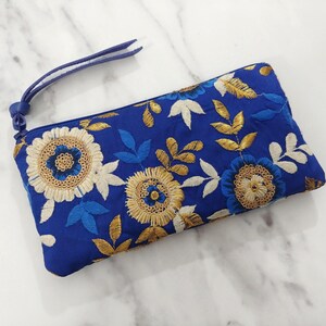 Op de afbeelding: Een blauwe geborduurde clutch met een polsbandje. De clutch heeft een bloemenontwerp met gouden, witte en blauwe draad. De bloemen zijn geborduurd op een blauwe stof.