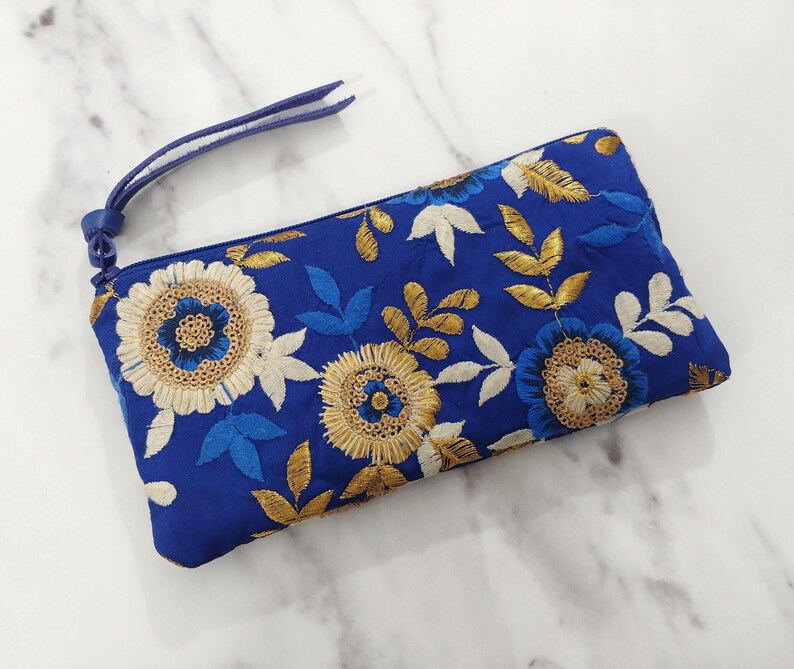 Op de afbeelding: Een blauwe geborduurde clutch met een polsbandje. De clutch heeft een bloemenontwerp met gouden, witte en blauwe draad. De bloemen zijn geborduurd op een blauwe stof.