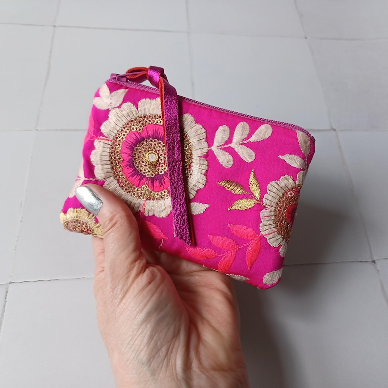 Op de afbeelding: Een felroze, geborduurde portemonnee met een bloemmotief. De portemonnee heeft cr&egrave;me- en gouddraad borduursel, een roze rits en een bijpassende kwast. De portemonnee wordt in een hand gehouden.