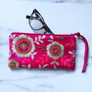 Op de afbeelding: Een felroze brillenkoker met bloemenborduurwerk en een bijpassende rits. De koker heeft een polsbandje en ligt naast een bril met schildpadmotief.
