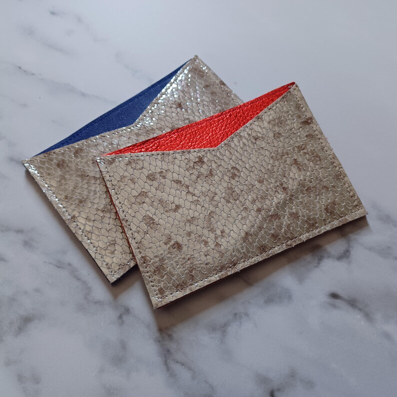 May include: Two leather cardholders with a snakeskin pattern. One has a blue interior and the other has a red interior. The cardholders are rectangular with a v-shaped opening.