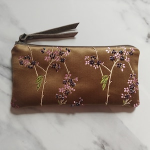 May include: A brown satin clutch with a zipper closure and a wrist strap. The clutch is embroidered with pink and purple floral designs and green leaves. The zipper and wrist strap are a matching brown color.