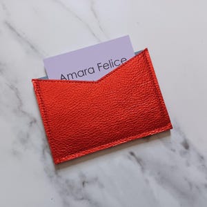 May include: A bright red leather card holder with the text "Amara Felice" on a light purple card. The card holder is rectangular with a unique angled top design. The card holder is made of textured leather.